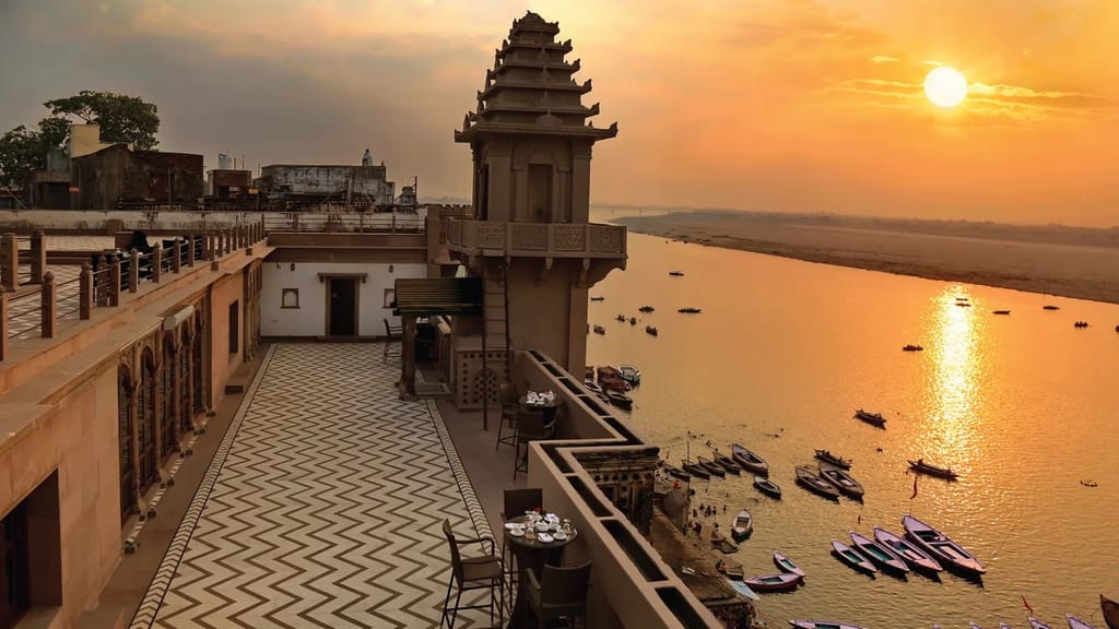 Sunrise over the Ganges from BrijRama Palace's rooftop terrace in Varanasi, zigzag marble floor and carved tower in the foreground, boats moored below