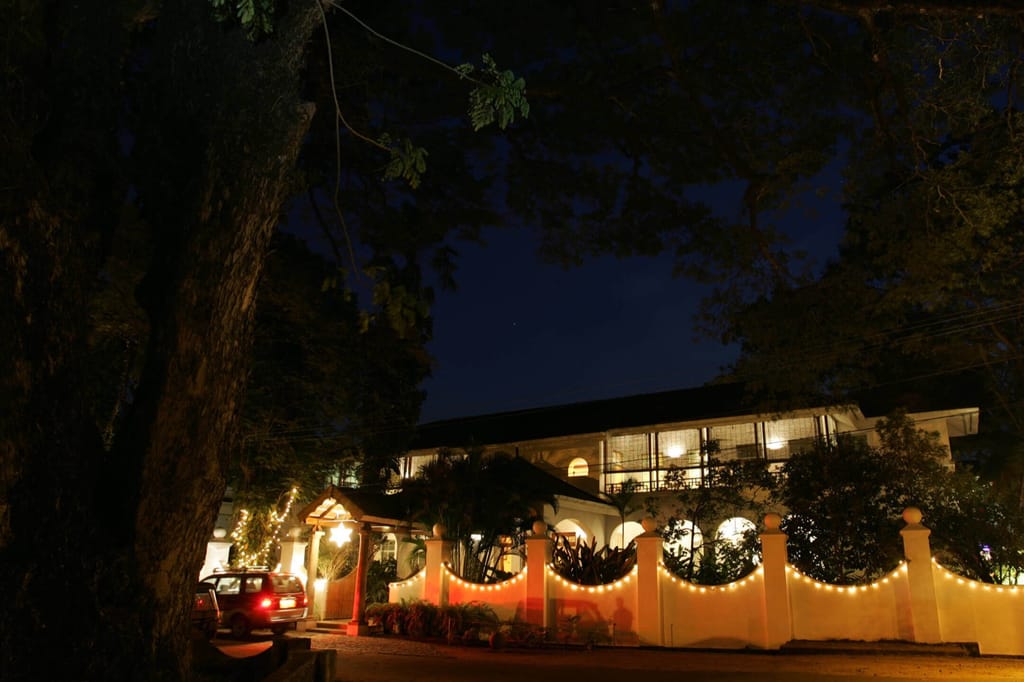 The Malabar House exterior at night, white gateposts and facade lit beneath old trees on Parade Road