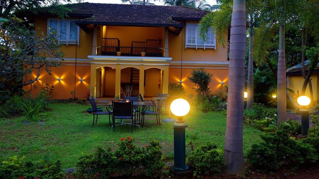 Tea Bungalow, Fort Kochi: the ochre two-storey heritage house at dusk, with garden lamps lit, first-floor balconies and iron tables on the lawn