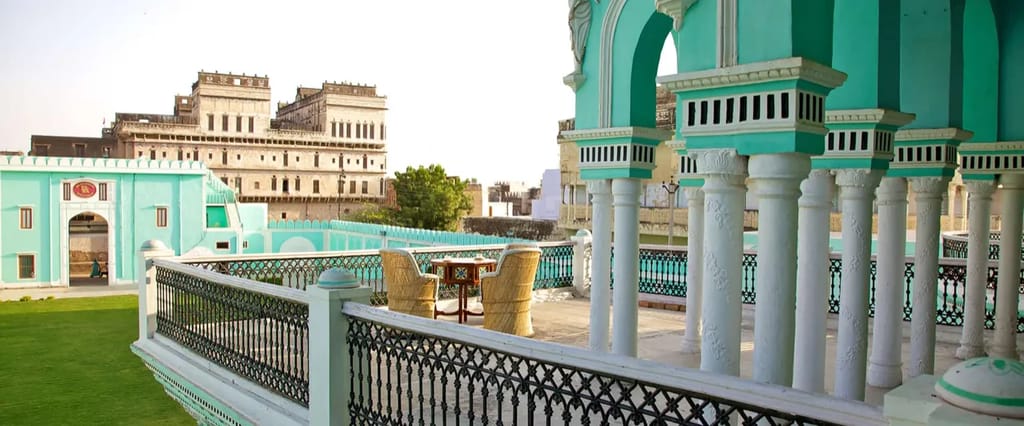 Rooftop terrace with aquamarine arched pavilion, carved white columns, black wrought-iron railings and cane chairs, looking across the lawn and gate to a large unrestored painted h
