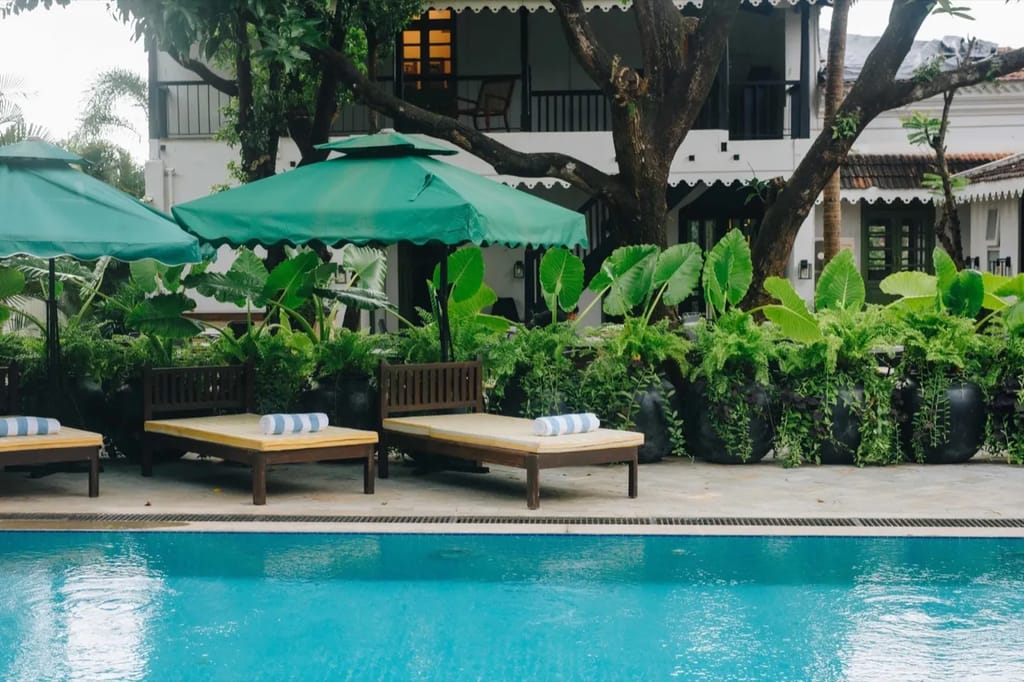 The pool at the Goa villa: turquoise water, yellow-striped sun loungers with rolled towels, green scalloped parasols, giant elephant-ear plants and the white two-storey villa with 
