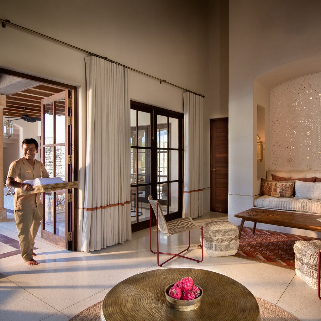 Kothi living room with cushioned seating nook, hammered brass table and staff member bringing towels at Aramness, Sasan Gir