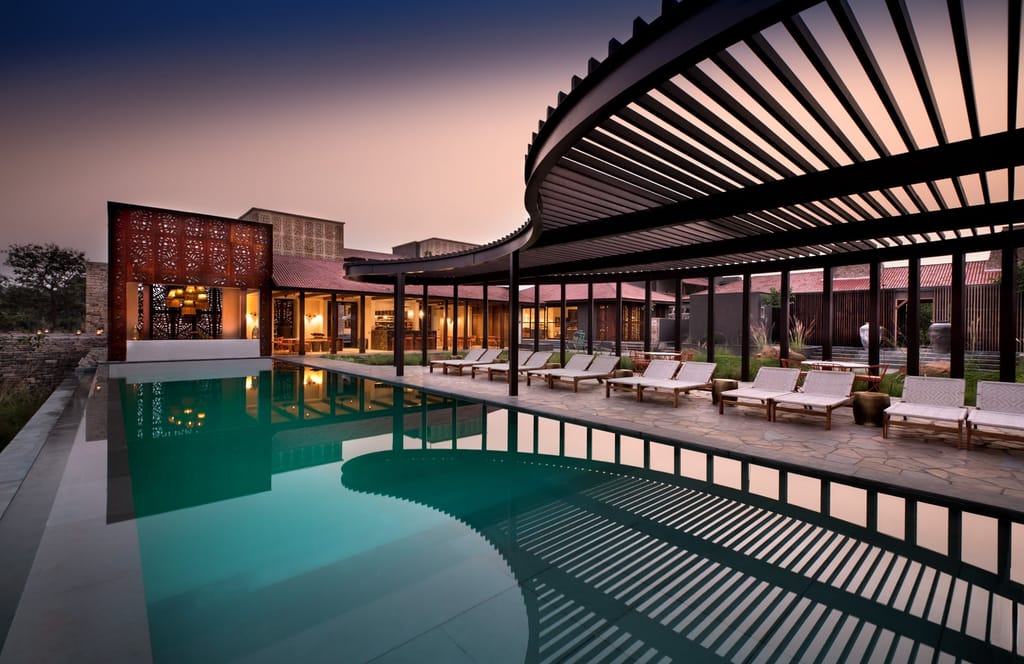 Main swimming pool with curved slatted pergola, loungers and lit lodge buildings at dusk at Aramness, Sasan Gir