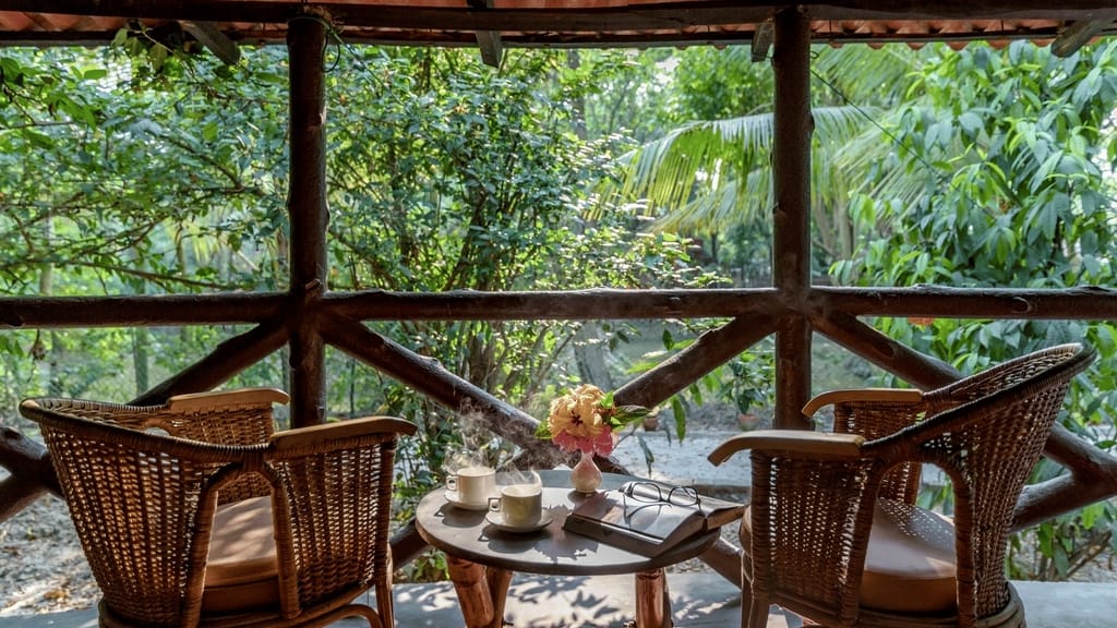 Executive Cottage — cane chairs and steaming coffee on the log-railed balcony facing greenery at Sundarban Tiger Camp, Sundarbans