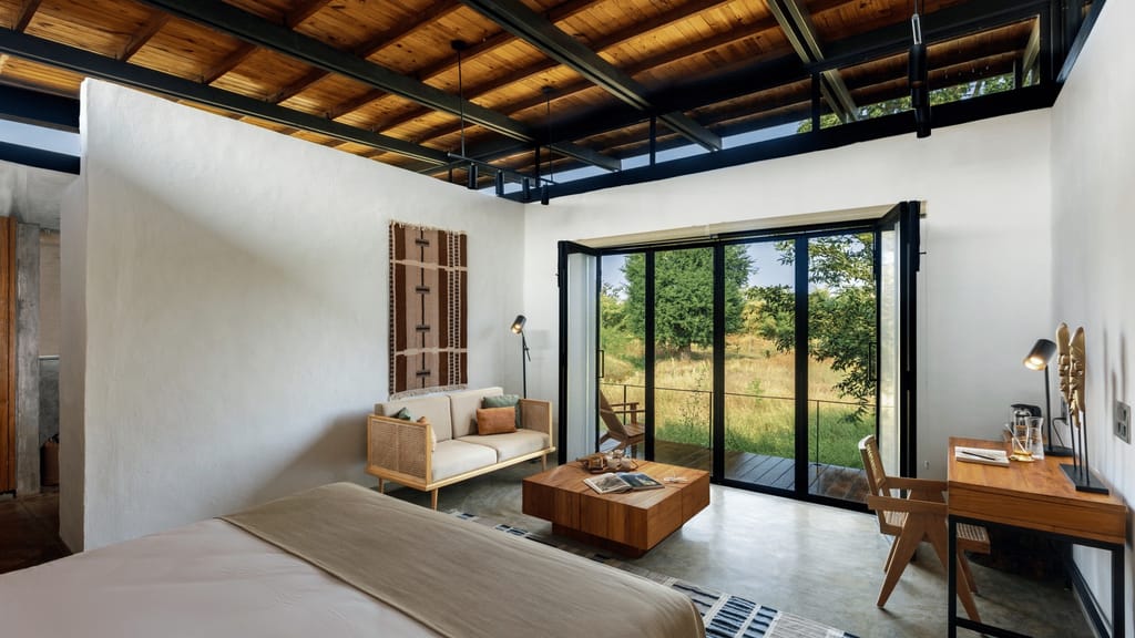 Luxury Room — bed and cane armchair seating beside folding glass doors opening to the grassland at Bori Safari Lodge, Satpura
