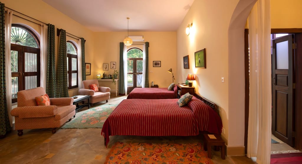 Bedroom with two beds in red striped covers, armchairs and arched windows at Mela Kothi – Chambal Safari Lodge, Chambal