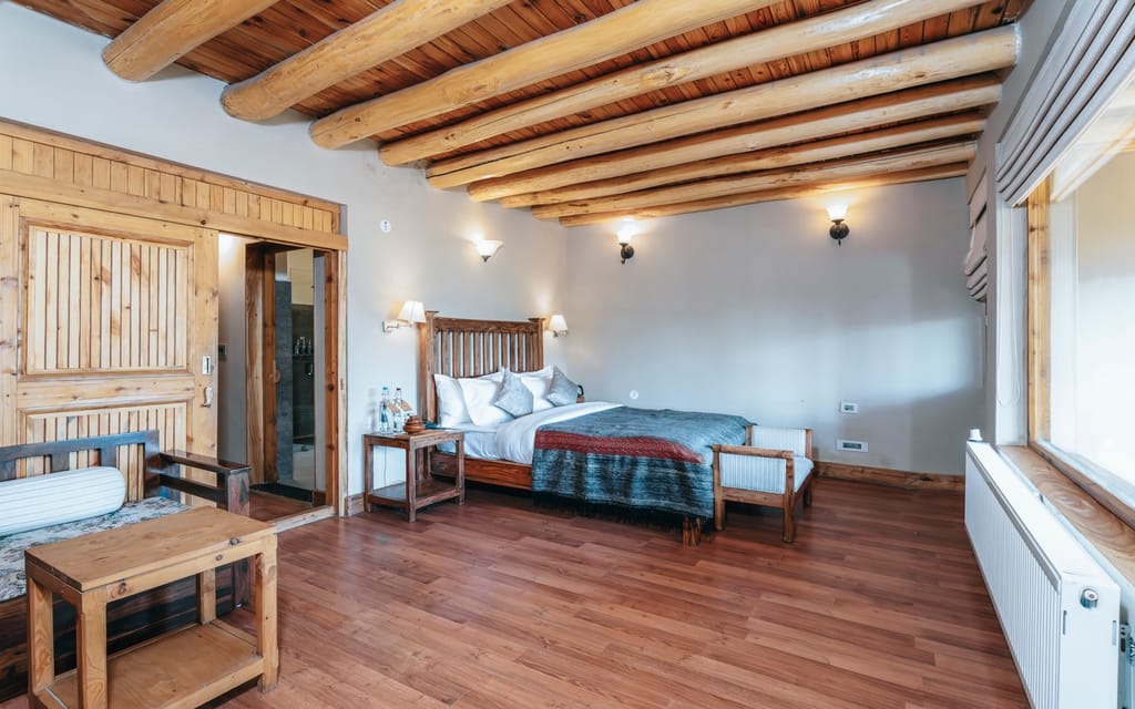 Duplex room with log-beamed ceiling, slatted wooden headboard, grey-and-red bed throw and wooden day bed at Ladakh Sarai, Leh