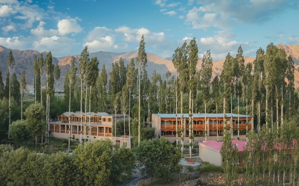 Mud-plastered guest wings and rooftop glasshouses among tall poplar trees, bare mountain slopes behind, at Ladakh Sarai, Leh