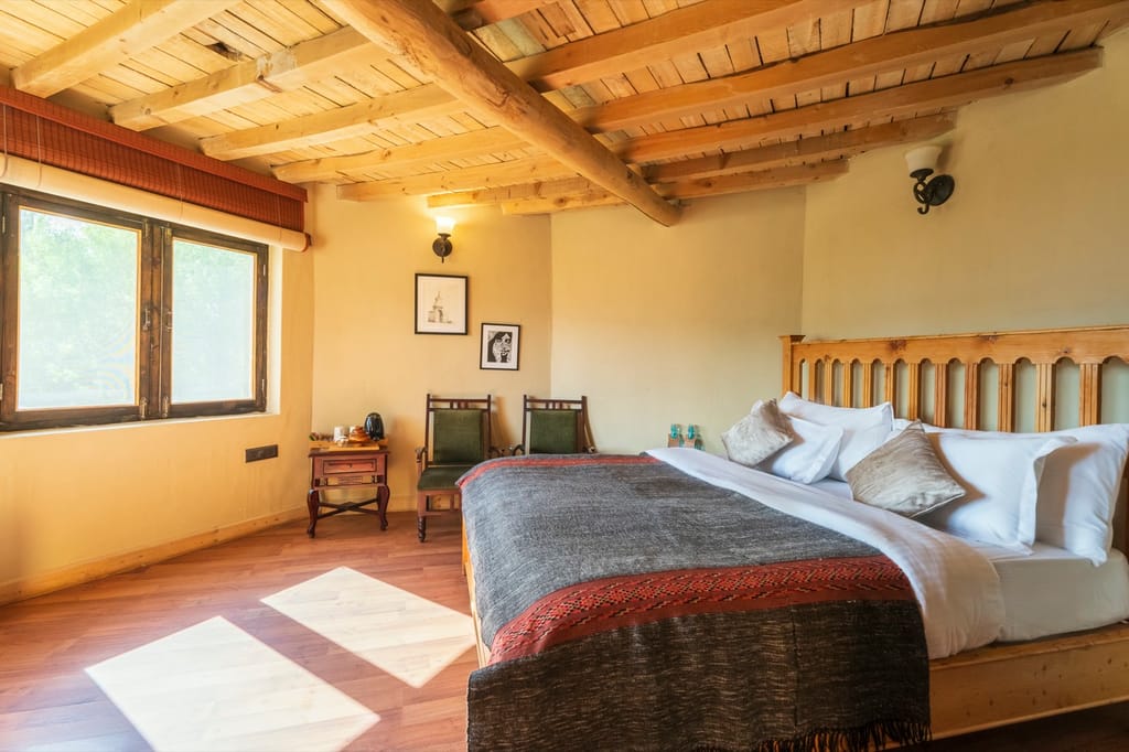 Mud Huts bedroom with timber ceiling, arched wooden headboard, woven grey throw and green chairs by the window at Ladakh Sarai, Leh