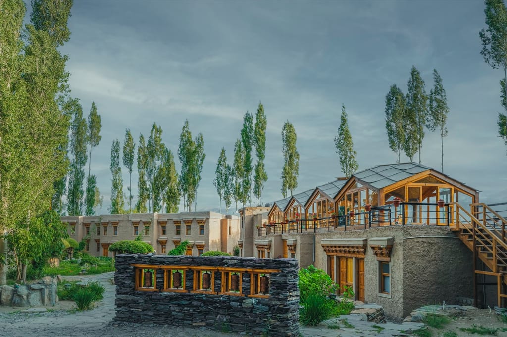 Stone wall of prayer wheels in the garden, with guest wings and a rooftop glasshouse terrace behind, at Ladakh Sarai, Leh