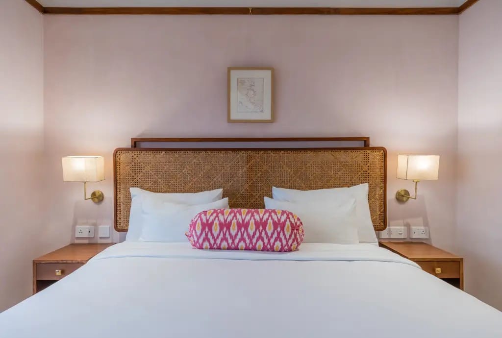 Pulaha double bed with woven cane headboard, brass wall lamps and soft pink walls at Amrit Bhawan, Haridwar
