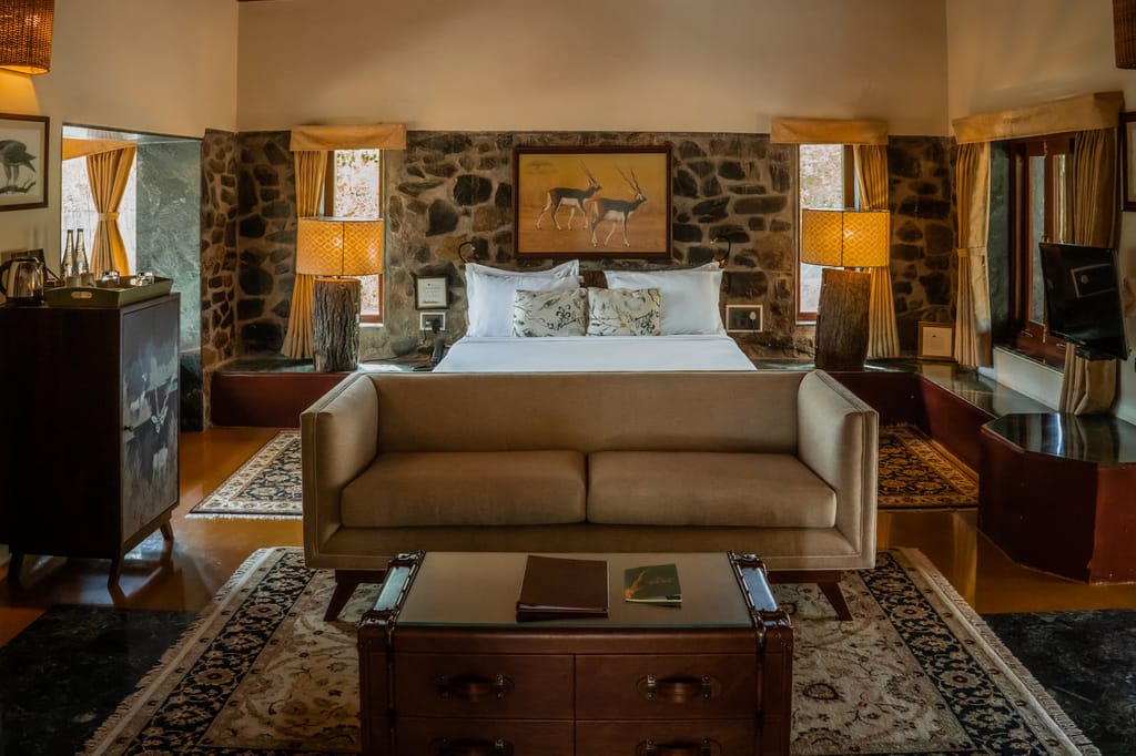 The Villa bedroom with stone walls, blackbuck painting above the bed, log-base lamps and beige sofa at The Blackbuck Lodge, Velavadar