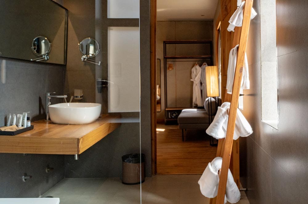 Club Room bathroom with white basin on a wooden counter and a towel ladder, looking through to the bedroom at The Chospa, Leh