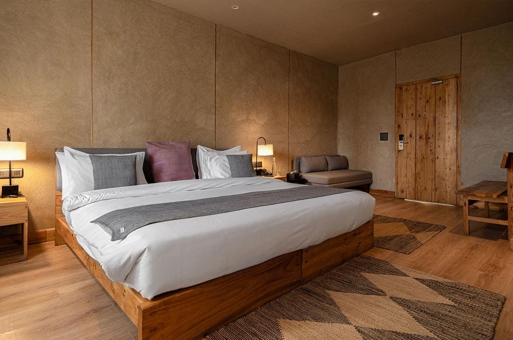 Club Room with low wooden platform bed, white linens, grey throw and hand-plastered earthen walls at The Chospa, Leh