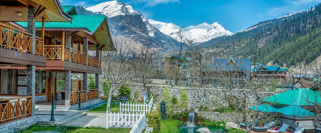 Wooden chalet balconies with green roofs overlooking gardens, a waterfall pond and snow-capped peaks at Storii Urvashi's Retreat, Manali