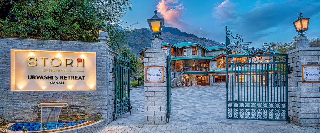 Stone gateposts and wrought-iron gates at the entrance, with the hotel and mountains behind at dusk, at Storii Urvashi's Retreat, Manali