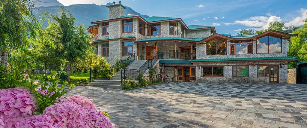 Stone-and-timber main building with green roofs, arched windows and a paved forecourt framed by hydrangeas at Storii Urvashi's Retreat, Manali
