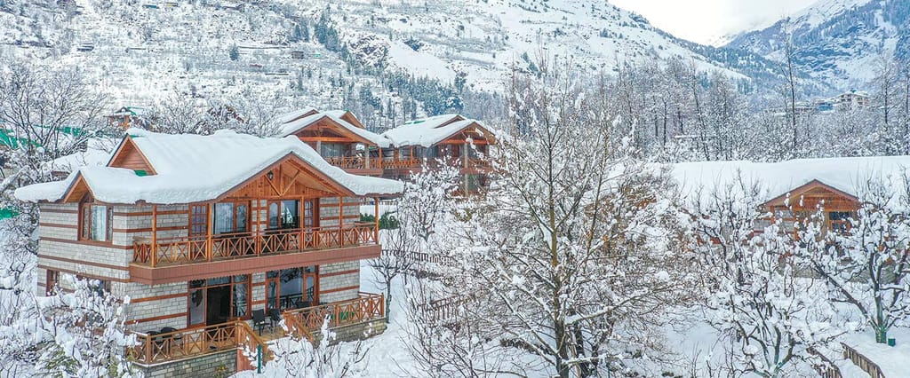 Snow-covered stone-and-wood chalets among bare trees on a white hillside in winter at Storii Urvashi's Retreat, Manali