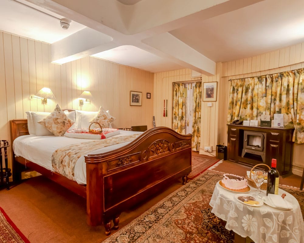 The Snuggery bedroom with carved wooden bed, panelled cream walls, floral curtains and a fireplace stove at Windamere Hotel, Darjeeling