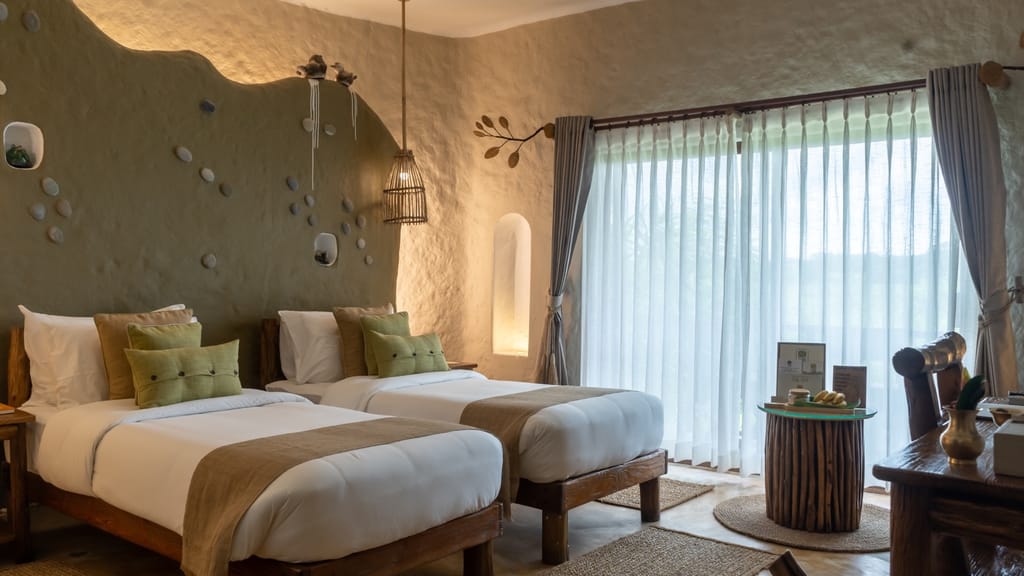 Twin beds with green cushions against a sculpted stone-studded wall and sheer curtains at Barahi Jungle Lodge, Chitwan