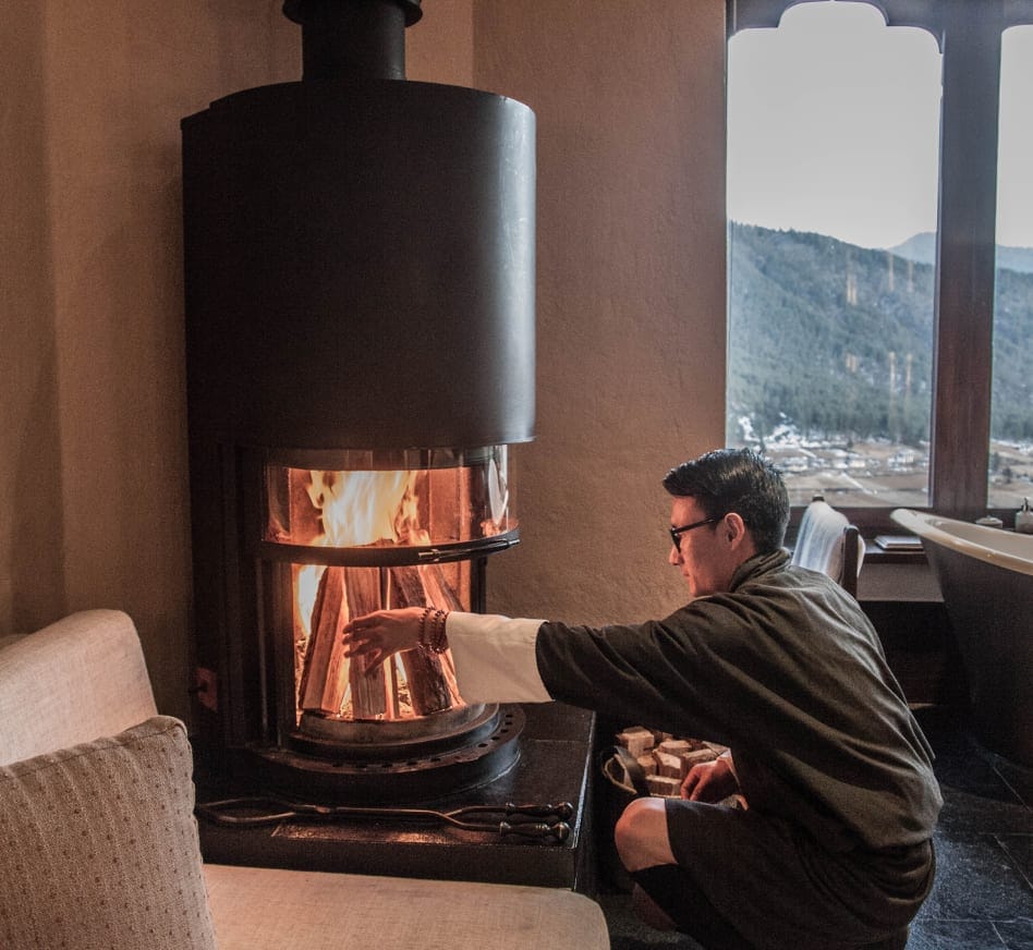 Bhutanese Farmhouse Suite staff member in traditional dress lighting the wood-burning stove at Gangtey Lodge, Gangtey