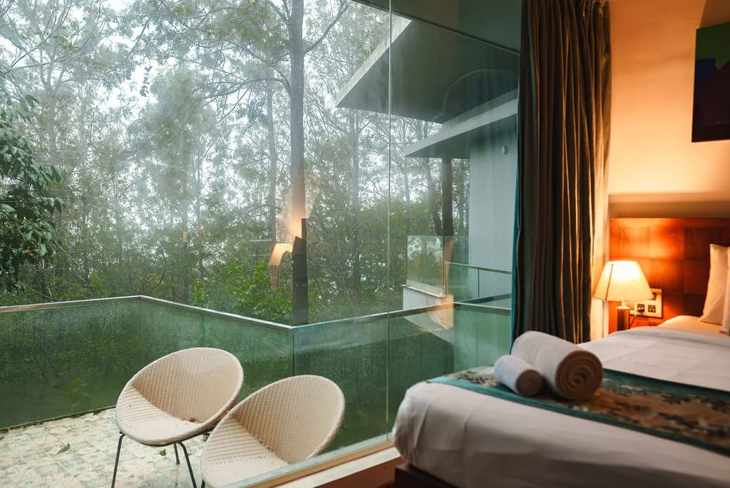 Qahva villa bedroom beside a glass-railed balcony with wicker chairs overlooking misty forest at Java Rain Resort, Chikmagalur
