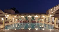 Courtyard swimming pool at dusk ringed by lit arches, curtained pavilions and corner chhatris