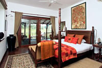 Villa bedroom with wooden four-poster bed, orange textiles and glass doors opening to a private deck at Samode Safari Lodge, Bandhavgarh