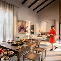 Kothi interior with brass-chained swing daybed, dining table laid for breakfast and staff member carrying a tray at Aramness, Sasan Gir