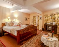 The Snuggery bedroom with carved wooden bed, panelled cream walls, floral curtains and a fireplace stove at Windamere Hotel, Darjeeling