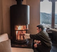 Bhutanese Farmhouse Suite staff member in traditional dress lighting the wood-burning stove at Gangtey Lodge, Gangtey