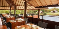 Open-sided restaurant with laid wooden tables looking across the lake to thatched dwellings at Jetwing Vil Uyana, Sigiriya
