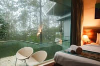 Qahva villa bedroom beside a glass-railed balcony with wicker chairs overlooking misty forest at Java Rain Resort, Chikmagalur