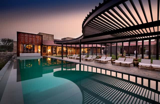 Main swimming pool with curved slatted pergola, loungers and lit lodge buildings at dusk at Aramness, Sasan Gir