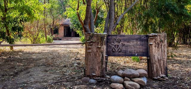 Carved wooden entrance sign on tree-trunk posts with a thatched cottage behind at Jim's Jungle Retreat, Corbett