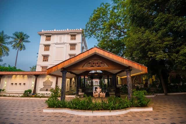 White multi-storey hotel building behind a tile-roofed entrance pavilion with garden beds at Svatma, Thanjavur