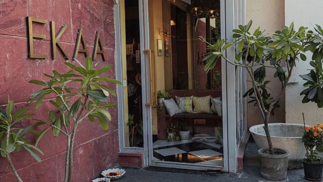 Entrance facade: red sandstone wall with brass EKAA lettering, glass door into a plant-filled lobby with chequerboard floor, frangipani trees in pots
