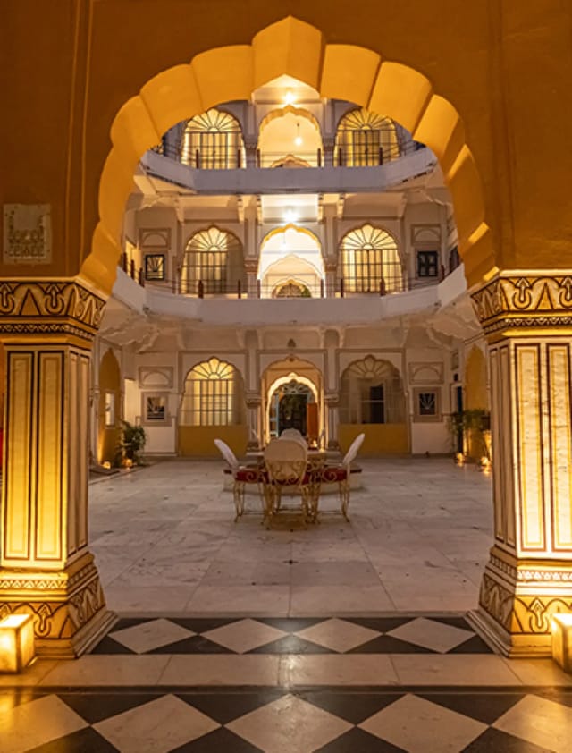 A lamplit inner courtyard of Hotel Diggi Palace seen through a scalloped arch at night, Jaipur