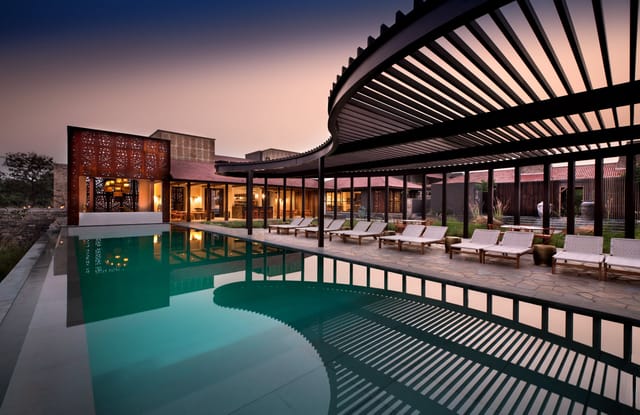 Main swimming pool with curved slatted pergola, loungers and lit lodge buildings at dusk at Aramness, Sasan Gir