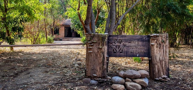 Carved wooden entrance sign on tree-trunk posts with a thatched cottage behind at Jim's Jungle Retreat, Corbett