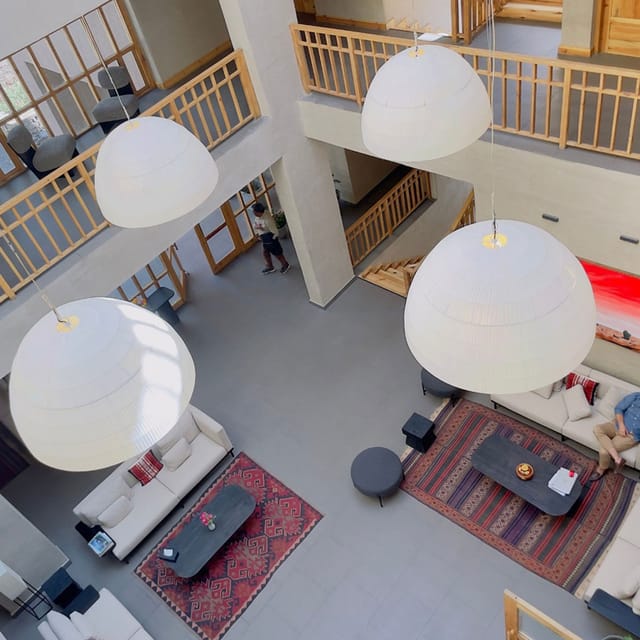 View from an upper walkway down into the double-height atrium lounge with oversized dome pendant lamps, sofas and red patterned rugs at The Chospa, Leh