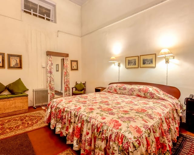 Ada Villa bedroom with high ceilings, rose-print bedspread and curtains, framed prints and Persian rugs at Windamere Hotel, Darjeeling