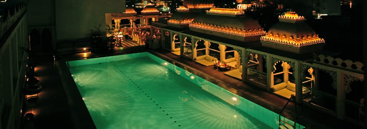 Rooftop swimming pool lit green at night beneath lamp-lined arcades and domed pavilions