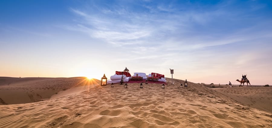 Private Dune Dinner table set on the sands at sunset with a camel approaching, Dhora Desert Resort & Spa, Shekhala