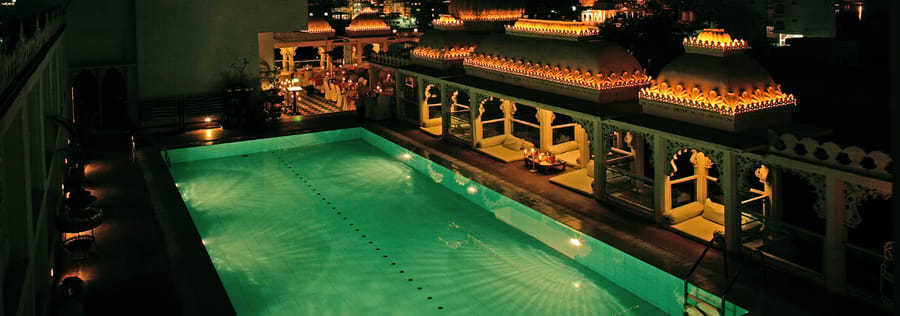 Rooftop swimming pool lit green at night beneath lamp-lined arcades and domed pavilions