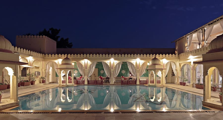 Courtyard swimming pool at dusk ringed by lit arches, curtained pavilions and corner chhatris