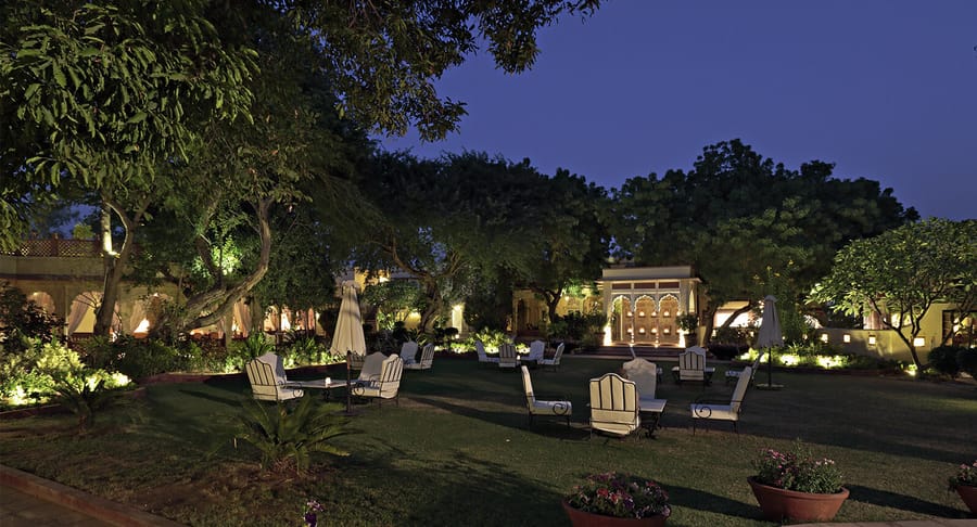 Lawns at night with lit trees, white garden chairs and the arcaded facade beyond