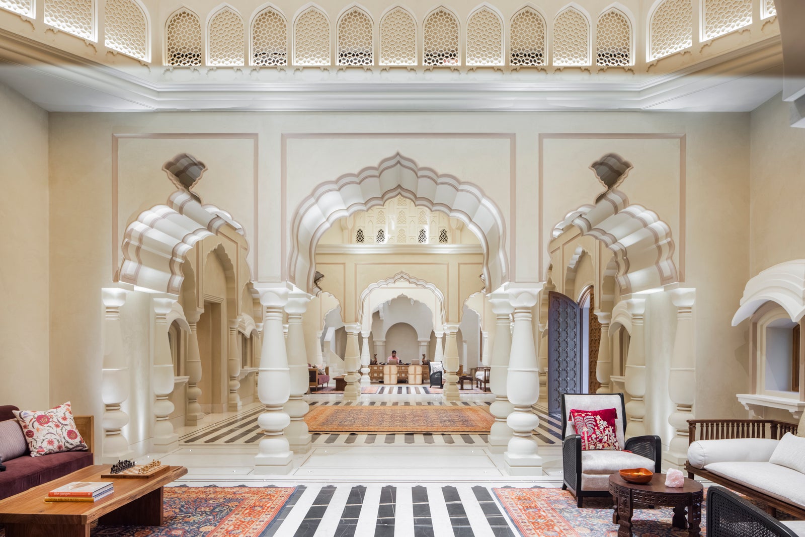 The lobby: scalloped Rajput arches, carved pillars, striped marble floor and layered rugs. One tiny distant figure at the desk, not identifiable.