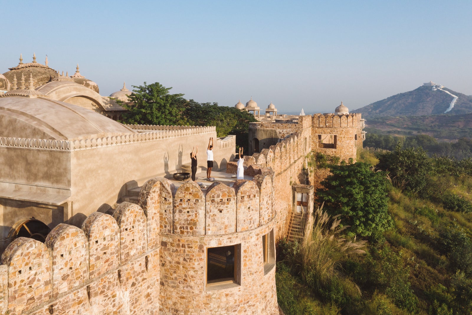 Sunrise yoga on a bastion of the fort wall, Chauth ka Barwara Mandir temple on the hill in the distance. Three small figures, backs turned, not identifiable.
