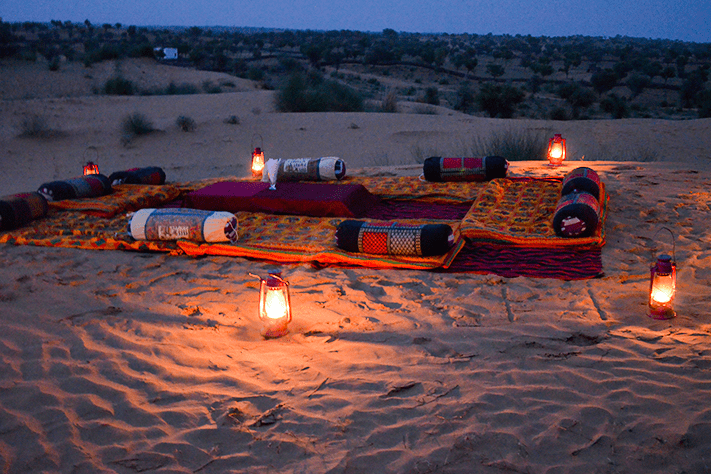 Desert dinner setup at dusk: floor seating with bolster cushions and rugs laid on sand dunes, ringed by lit hurricane lanterns, no people